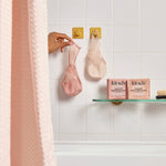 Person hanging a pink Kitsch shampoo bar in a Kitsch saver bag on a hook with Kitsch products on a bathroom shelf.