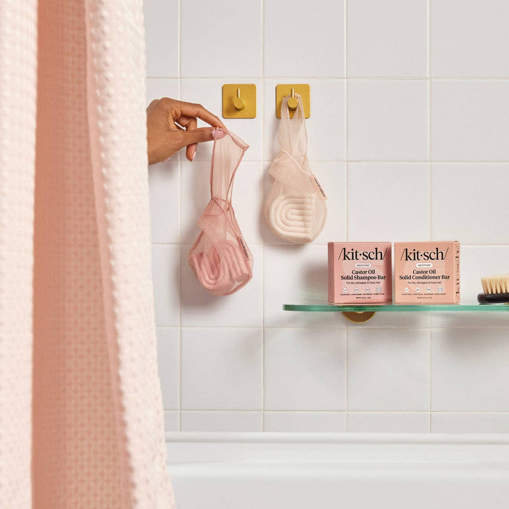 Person hanging a pink Kitsch shampoo bar in a Kitsch saver bag on a hook with Kitsch products on a bathroom shelf.