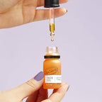 Hand holding a bottle of UpCircle Face Oil with a dropper of oil against a light purple background