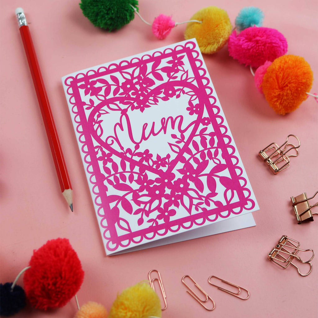 Card with 'Mum' design on pink background with leaves and flowers design. A red pencil, colourful pompoms and clips in the background.