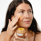 Woman applying yellow cleansing balm to her face with a jar of balm in hand on a white background