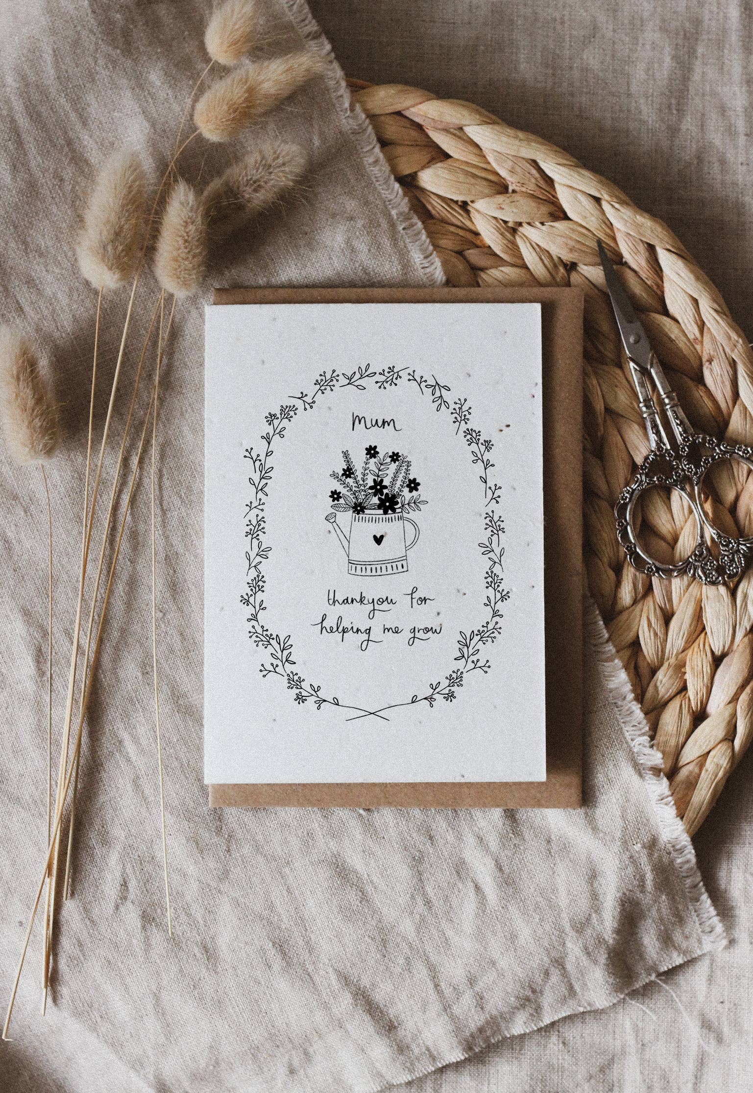Mother's Day Greeting card with floral design and text. Printed on seed paper. Placed on a textured surface with dried plants and scissors.