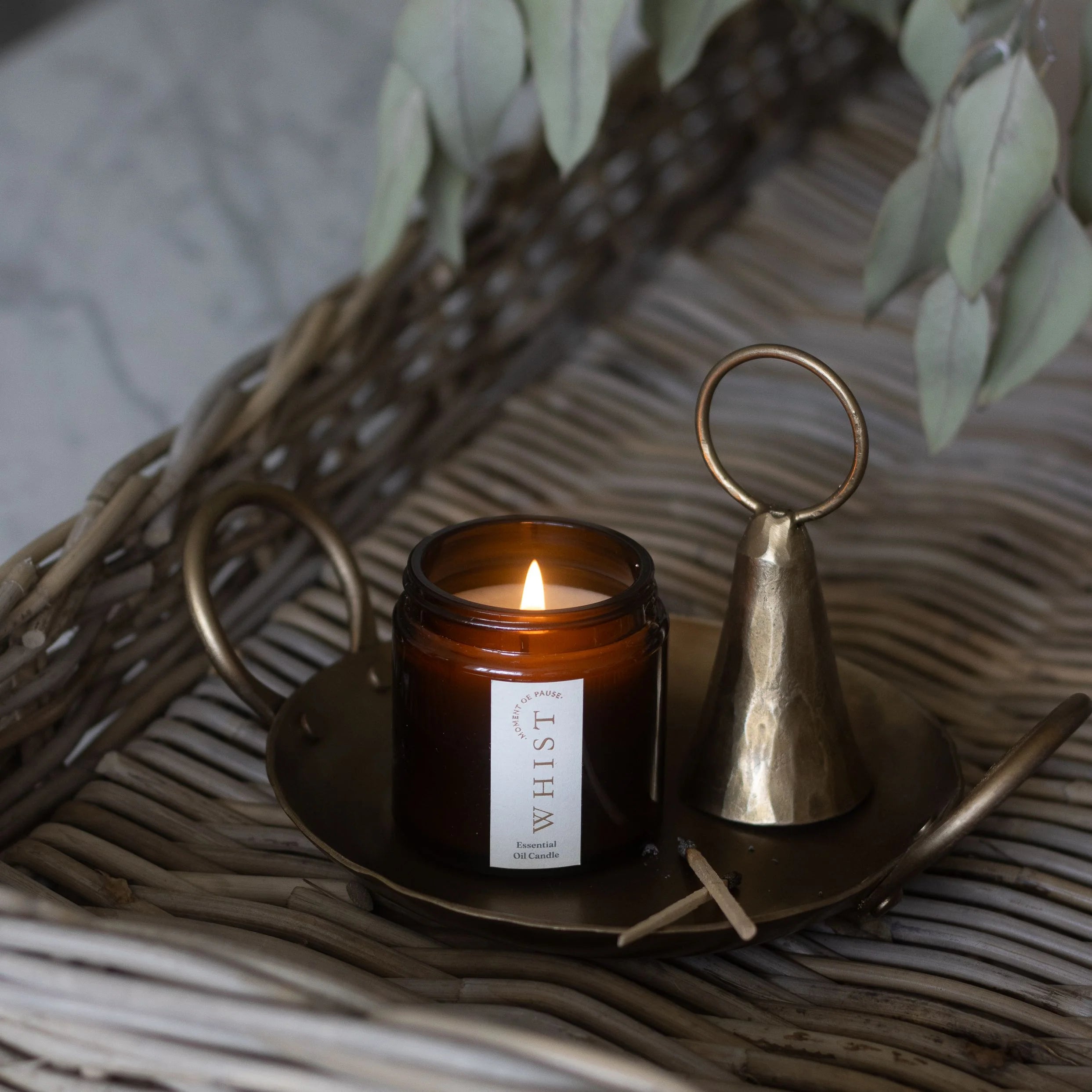 Lit candle in a brown amber jar with a visible Whist brand label, placed on a woven surface with a brass plate, brass candle snuffer, burnt matches and dried plant leaves.