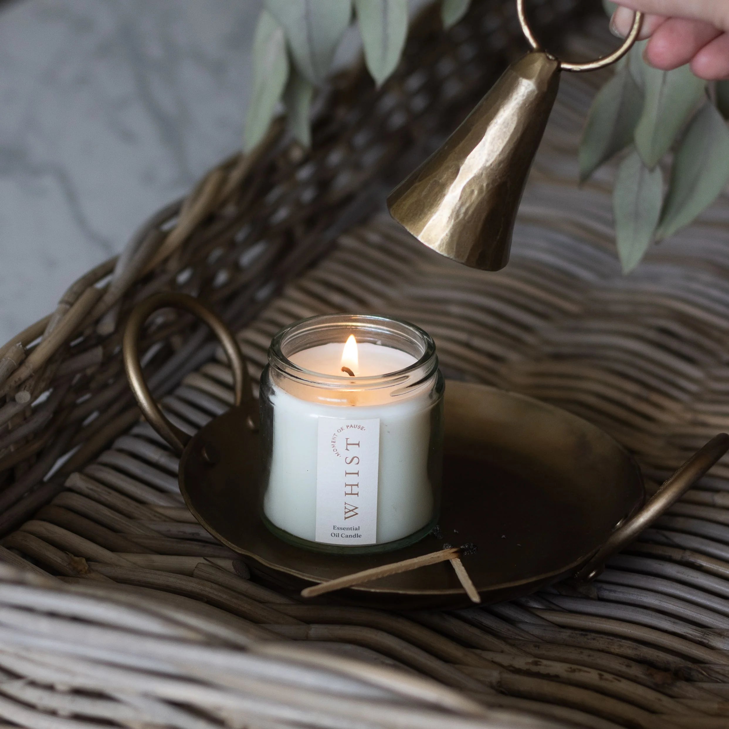 Candle in a glass jar with a visible Whist brand label, placed on a brass plate and a woven surface, with a brass candle snuffer. Used matches sit on the brass plate. Dried leaves are in the background.