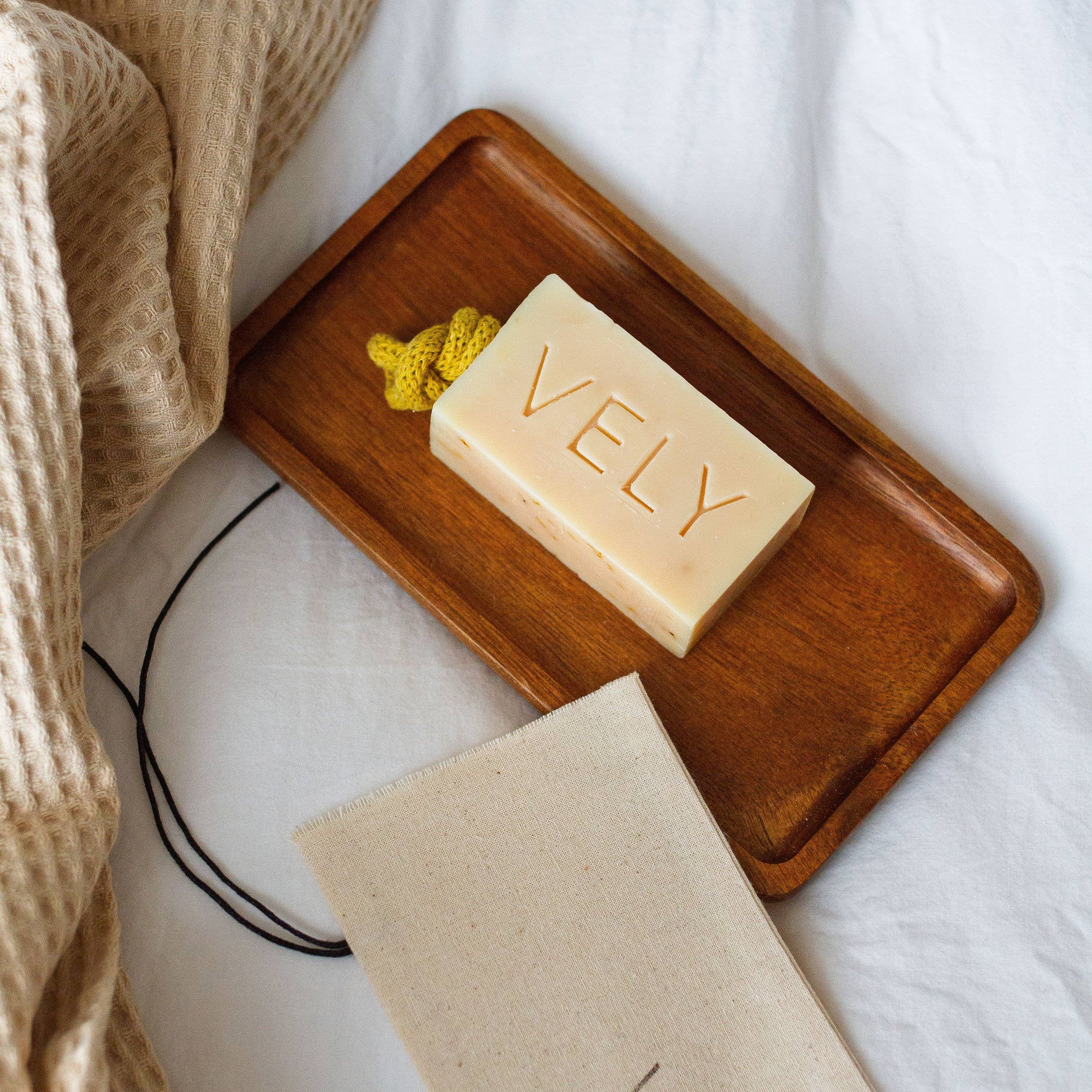 Bar of VELY soap on a wooden tray with a matching soap bag on a white background