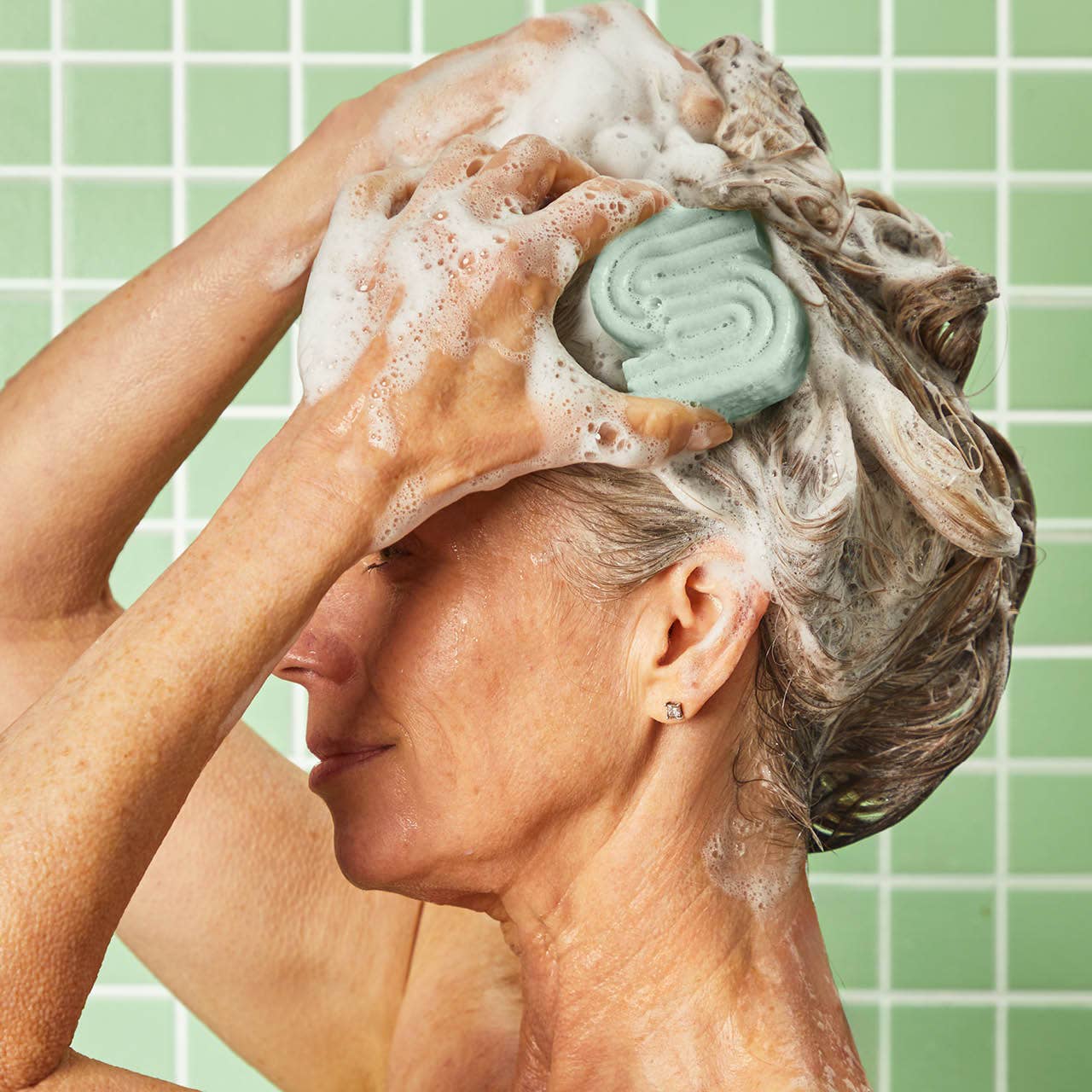 Woman washing her hair with a Kitsch Rosemary Volumising Shampoo Bar, against a green tiled wall.