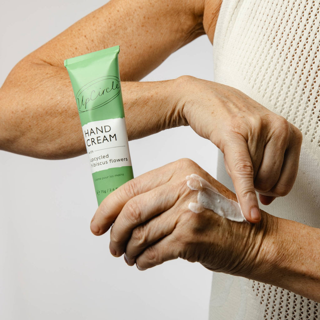 Person applying hand cream to their hand with a green tube labeled 'UpCircle Hand Cream' in the foreground.
