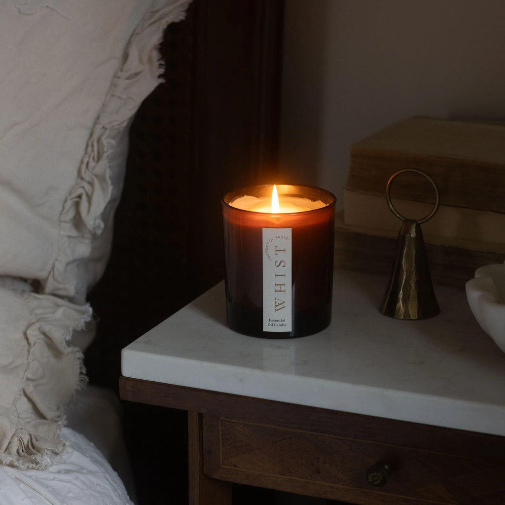 Lit candle in an amber glass jar, with a visible 'Whist' brand label on a nightstand next to a bed. In the background is a brass candle snuffer and vintage books.