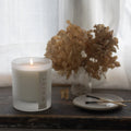 Lit candle in a frosted glass container and a 'Whist' brand label. Sitting on a wooden surface with dried flowers, and matches with a white curtain in the background