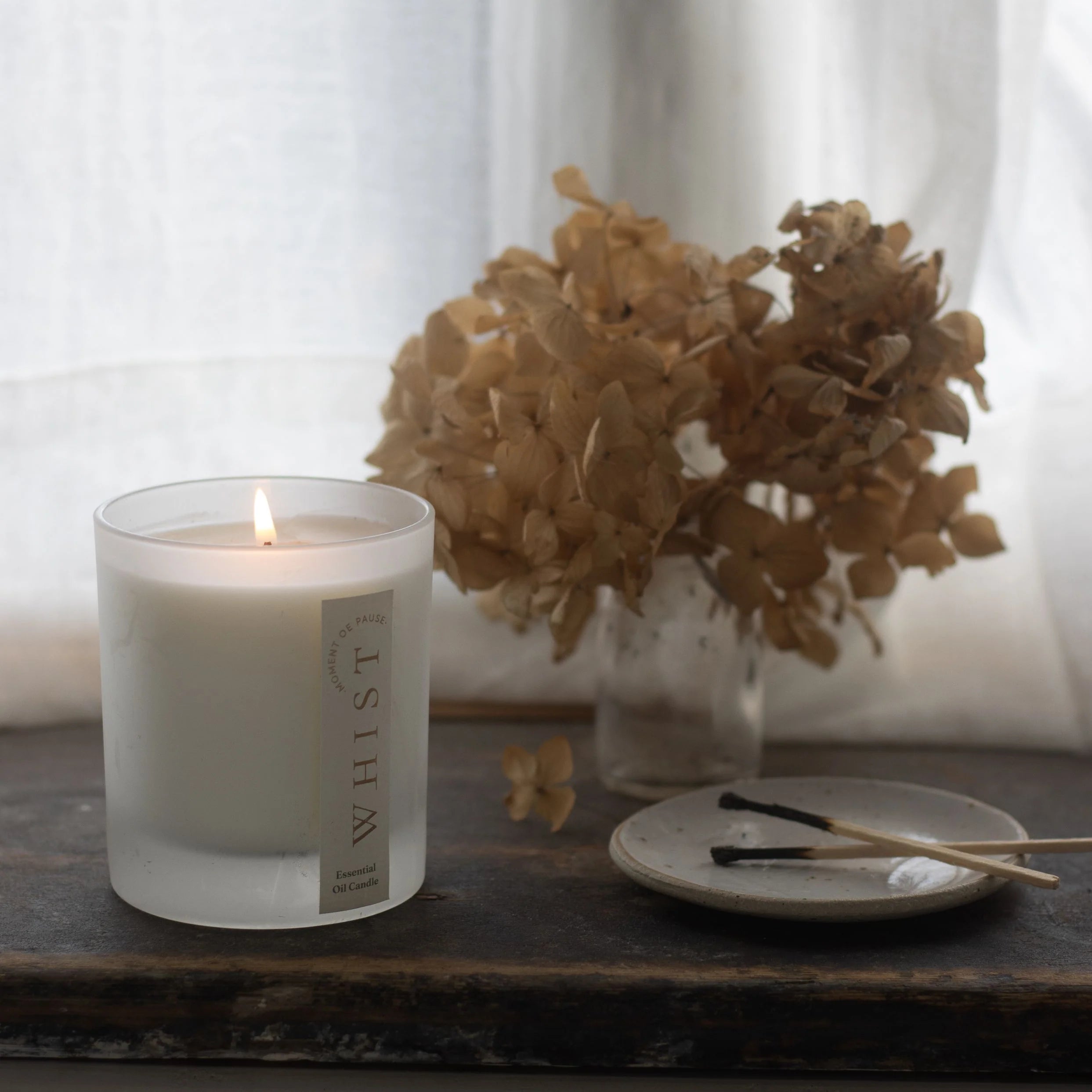 Lit candle in a frosted glass container and a 'Whist' brand label. Sitting on a wooden surface with dried flowers, and matches with a white curtain in the background