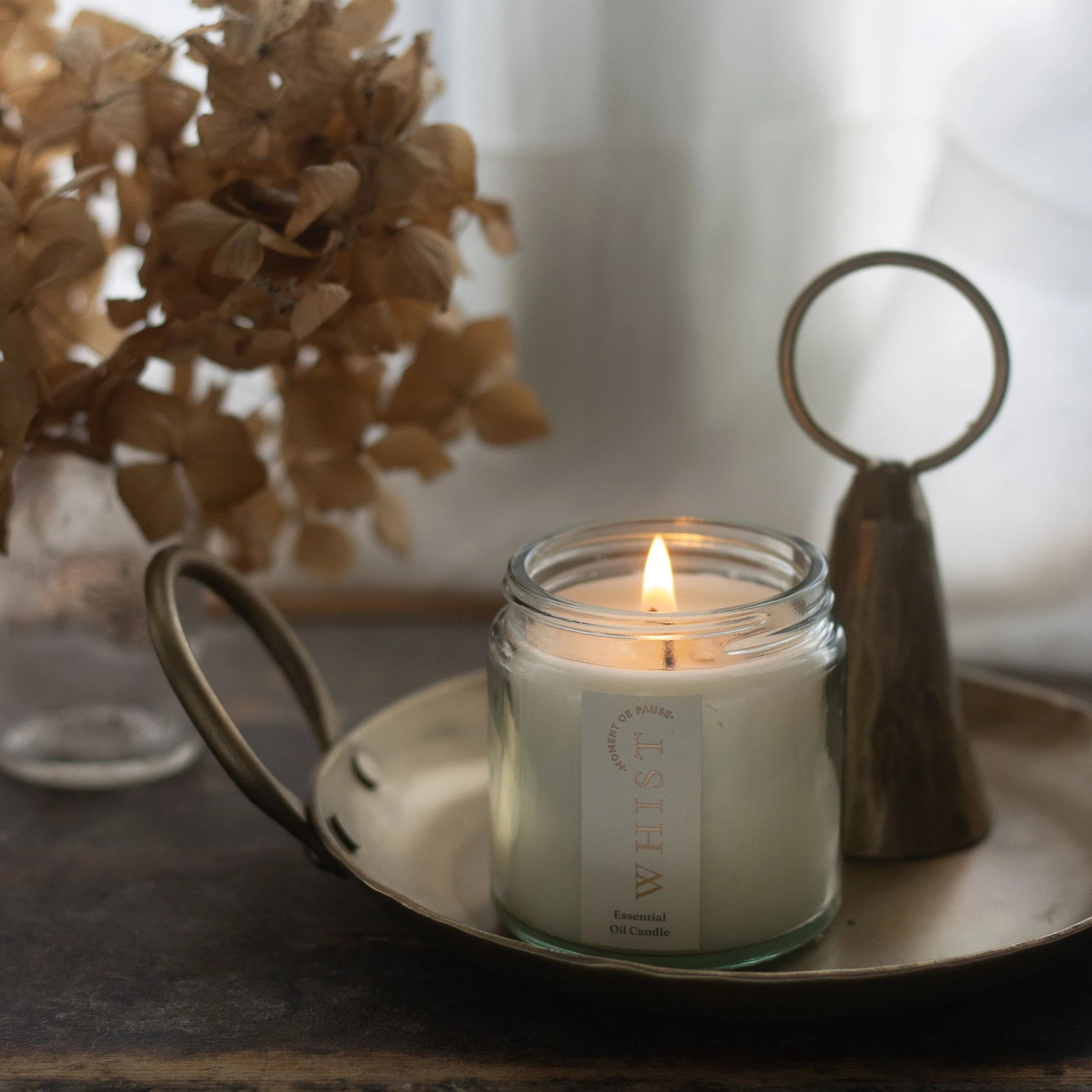Lit candle in a glass jar on a metal tray with decorative elements and dried flowers in the background. Set against a neutral background and on a wooden table