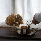 Person wearing a white top lighting a candle in an amber jar sitting on a brass plate with a brass candle snuffer. Dried flowers and a white curtain in the background