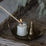 White candle in a clear glass jar and visible Whist branded label being lit with a wooden match. Sitting on a brass plate with a used match and brass candle snuffer. All items sit on a woven surface with dried greenery in the background.