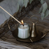 White candle in a clear glass jar and visible Whist branded label being lit with a wooden match. Sitting on a brass plate with a used match and brass candle snuffer. All items sit on a woven surface with dried greenery in the background.