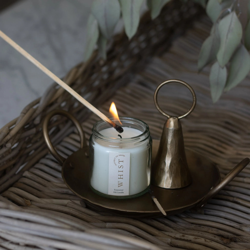 White candle in a clear glass jar and visible Whist branded label being lit with a wooden match. Sitting on a brass plate with a used match and brass candle snuffer. All items sit on a woven surface with dried greenery in the background.