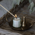 White candle in a clear glass jar and visible Whist branded label being lit with a wooden match. Sitting on a brass plate with a used match and brass candle snuffer. All items sit on a woven surface with dried greenery in the background.