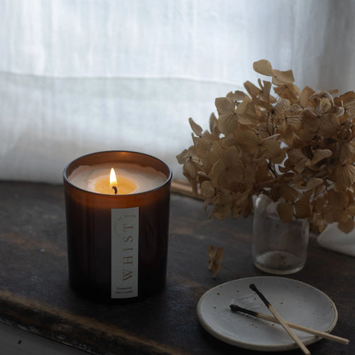 Lit candle in an amber glass jar with a visible 'Whist' brand label. On a wooden surface next to dried flowers and burnt matches on a plate.