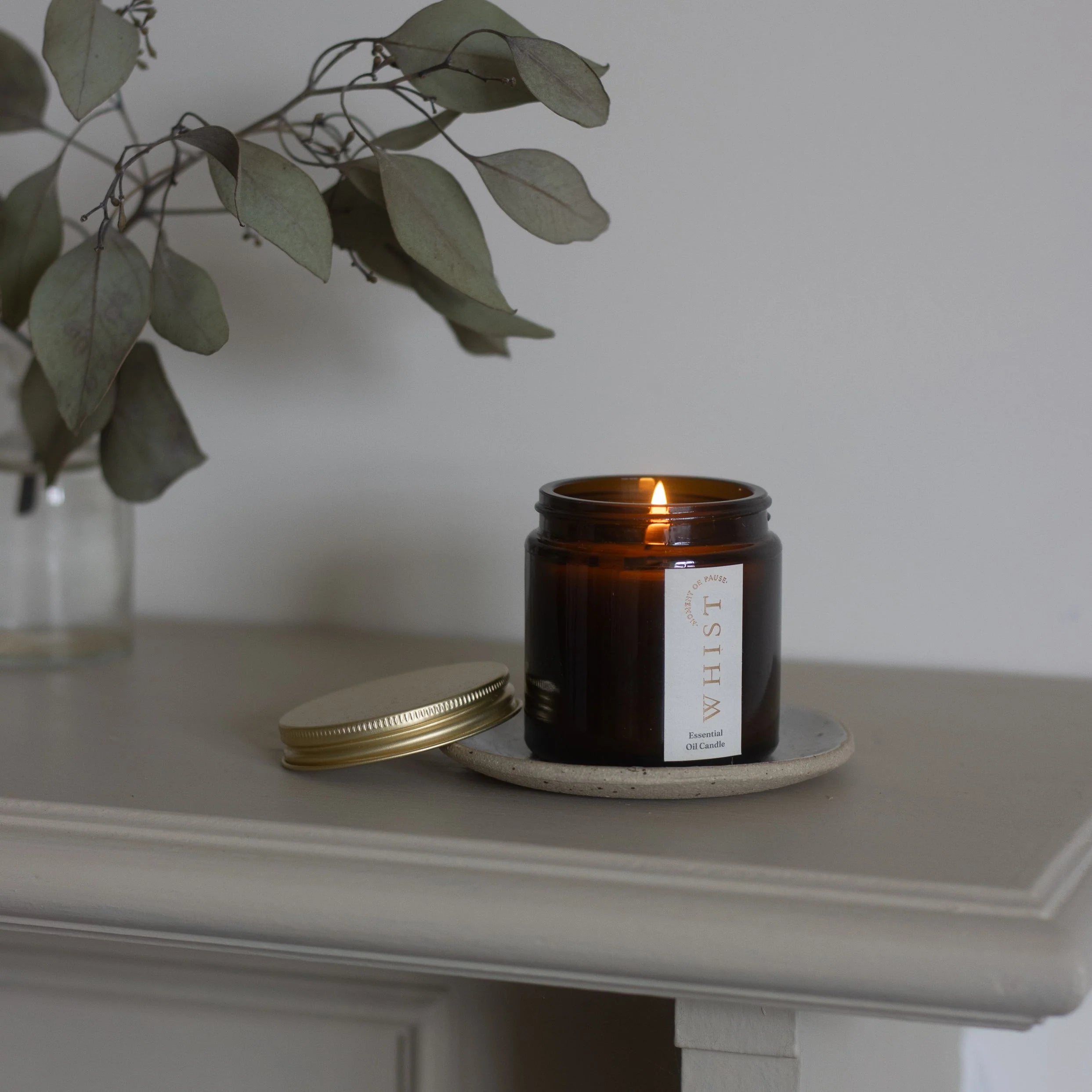 Lit candle in an amber glass jar and a gold lid with a visible Whist brand label. Sitting on a light surface, with a plant in the background.