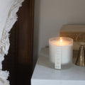 Lit candle in a frosted glass container with 'Whist' branding. On a marble bedside table with a brass candle snuffer and vintage books in the background.