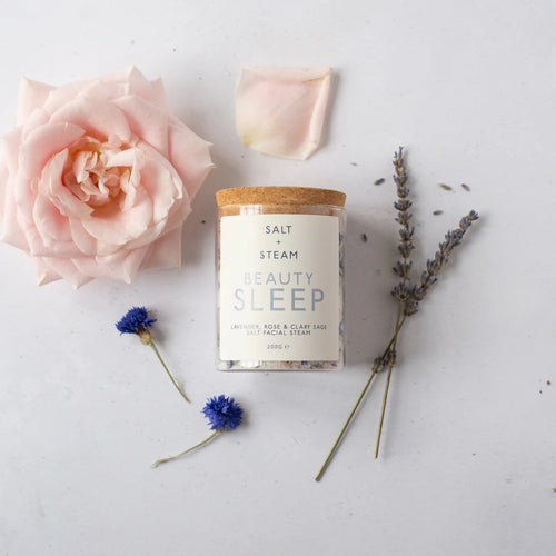 Glass Jar of Facial Steam salts with a cork lid, labeled 'Salt + Steam Beauty Sleep' with rose, lavender and cornflower, on a light background