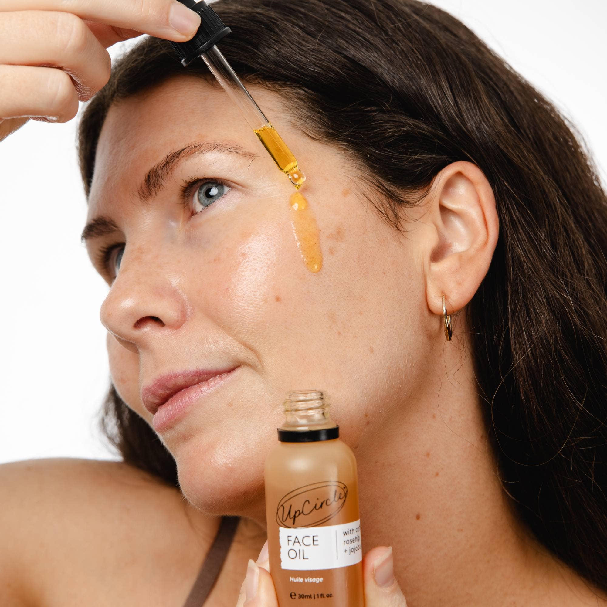 Woman applying face oil to her face with a dropper, holding a bottle labeled 'UpCycled Face Oil'.