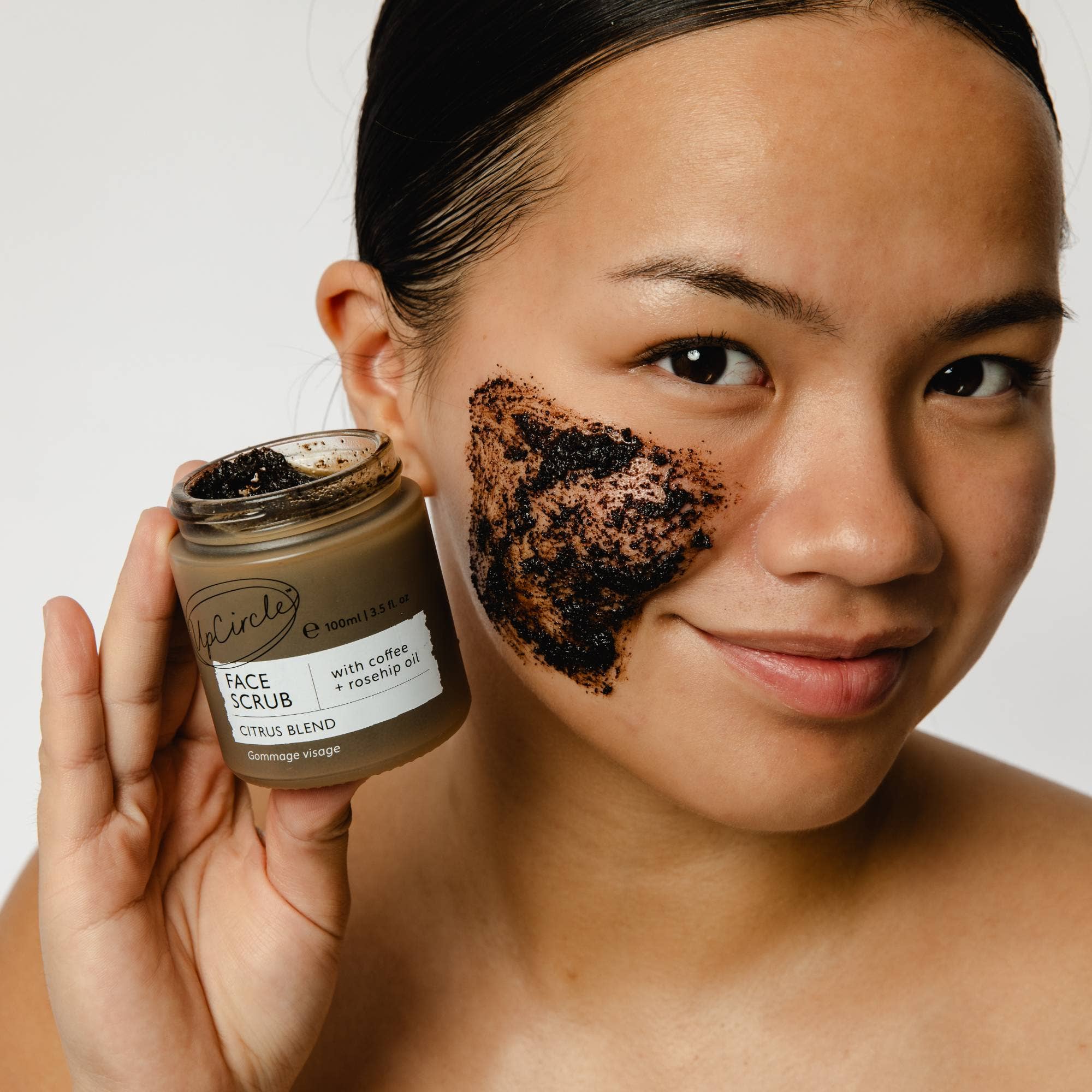 Woman applying coffee scrub to her face with a jar of Upcircle coffee and rosehip face scrub